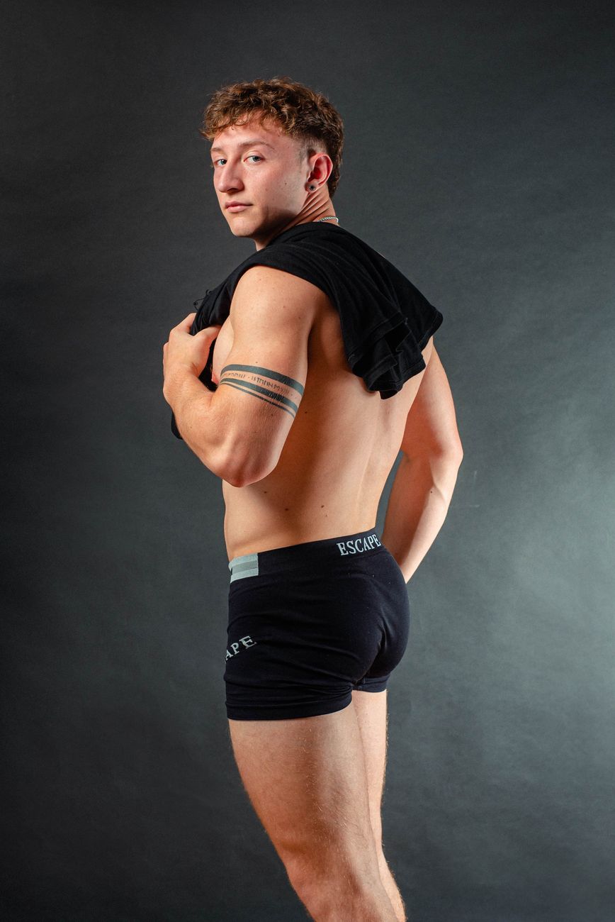 Confident male model posing in black boxer shorts during studio fashion shoot with a gray background.